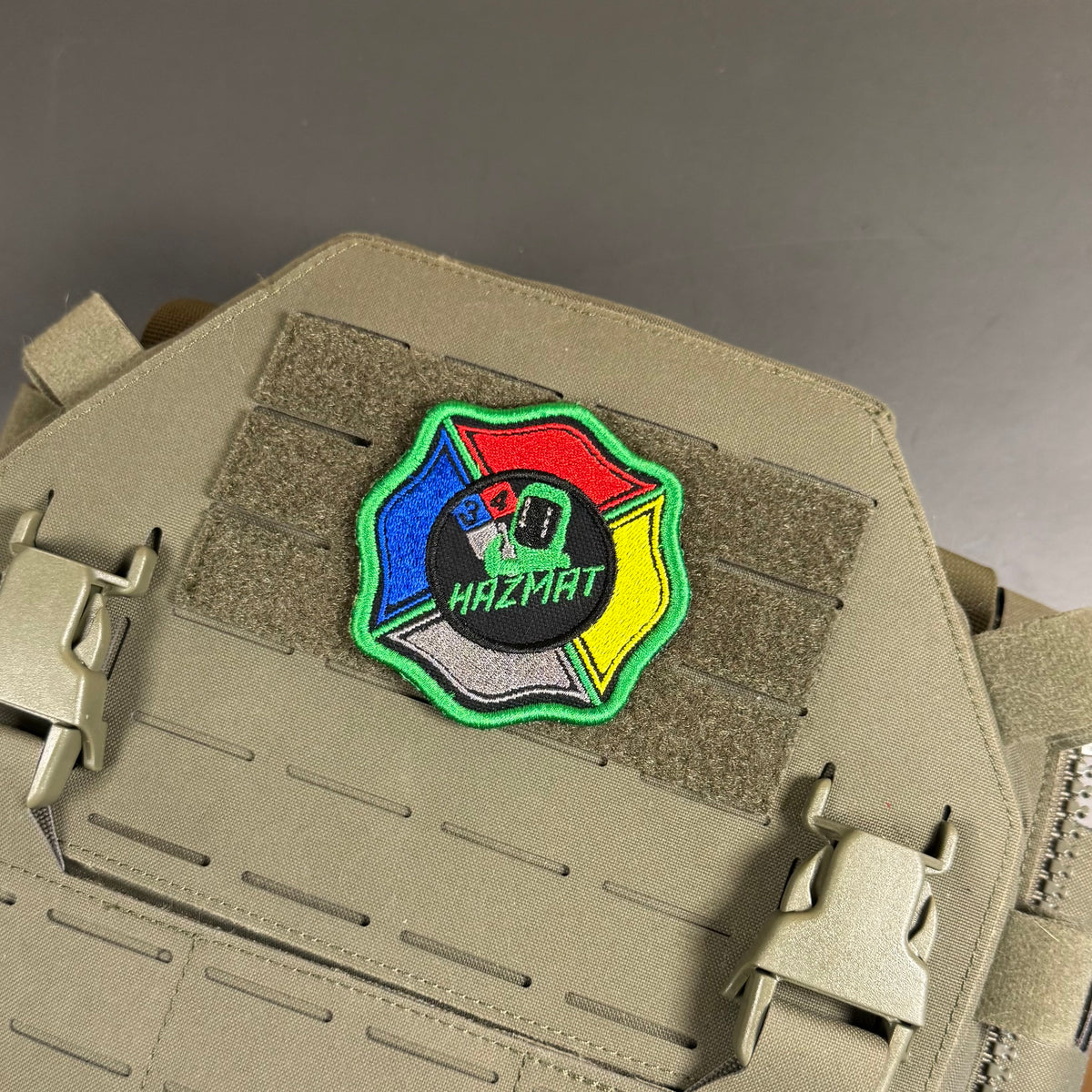 An embroidered Hazmat patch with a multicolor design (red, blue, yellow, black, green, white) mounted on the tan MOLLE webbing of a tactical vest.