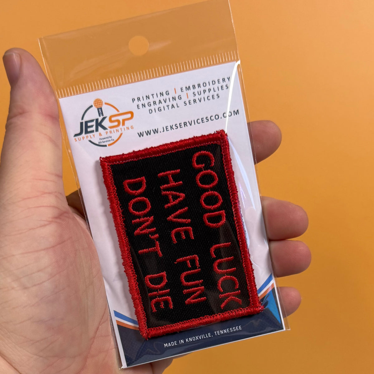 A hand holds a packaged embroidered patch that reads “GOOD LUCK HAVE FUN DON’T DIE” in red lettering on a black background, with a white backing card branded JEKSP Supplies & Services.