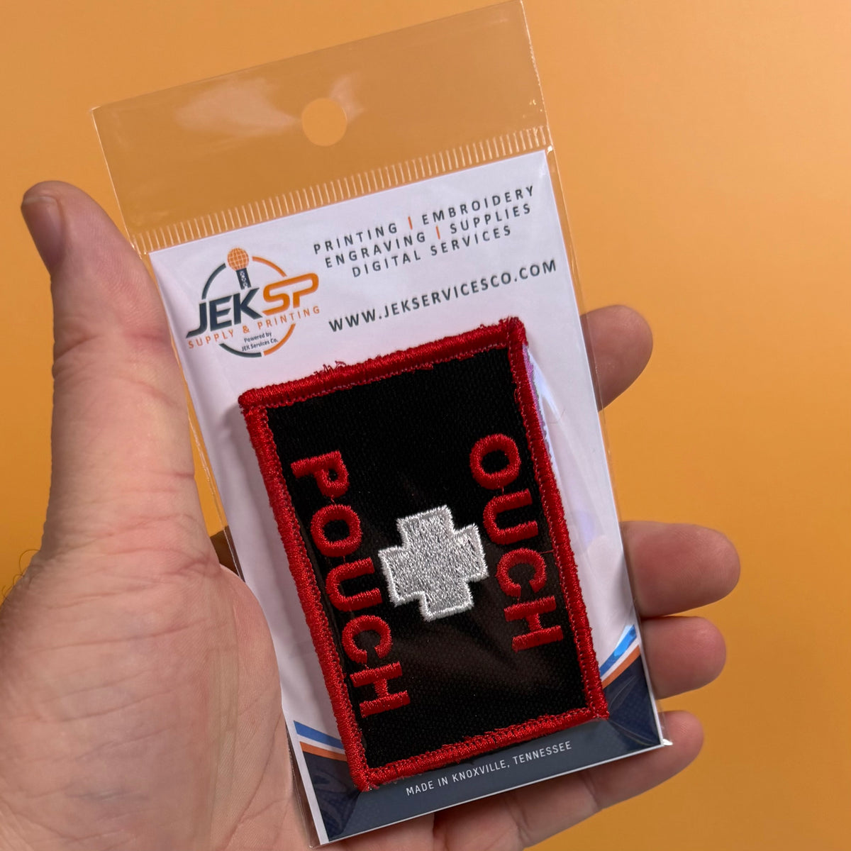 A hand holding a packaged embroidered patch that reads 'POUCH' in red with a silver cross on a black background, against an orange backdrop. The packaging card shows the JEKSP brand.