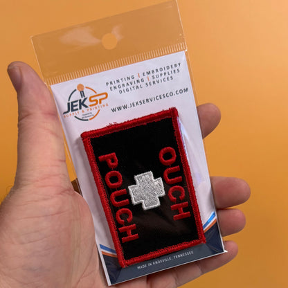 A hand holding a packaged embroidered patch that reads 'POUCH' in red with a silver cross on a black background, against an orange backdrop. The packaging card shows the JEKSP brand.