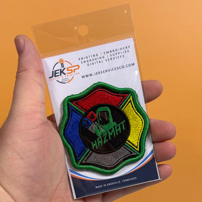 A hand holds a Jeksp embroidered patch in its original retail packaging against a solid orange background. The patch is a Maltese cross shape with the word 'HAZMAT' embroidered in green on a black center, surrounded by red, blue, yellow, and gray embroidered sections with a green border.