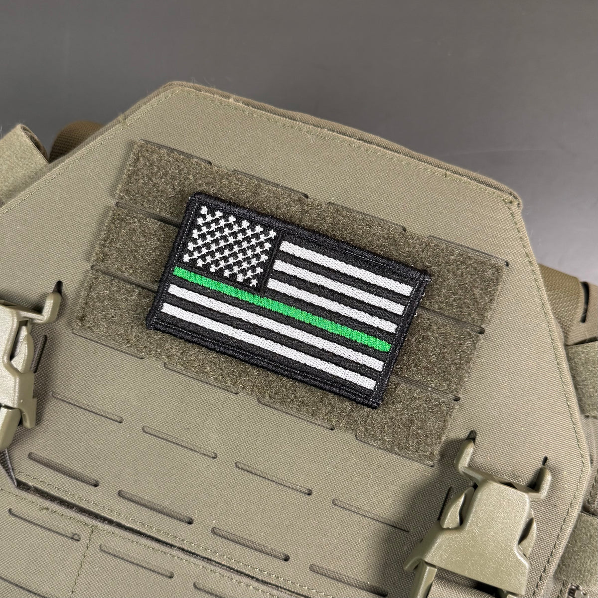 Thin Green Line Tactical American Flag embroidered morale patch: featuring a black-and-white American flag with a single green stripe, mounted on tan tactical gear with Velcro and plastic clips.