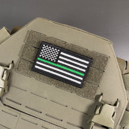 Thin Green Line Tactical American Flag embroidered morale patch: featuring a black-and-white American flag with a single green stripe, mounted on tan tactical gear with Velcro and plastic clips.