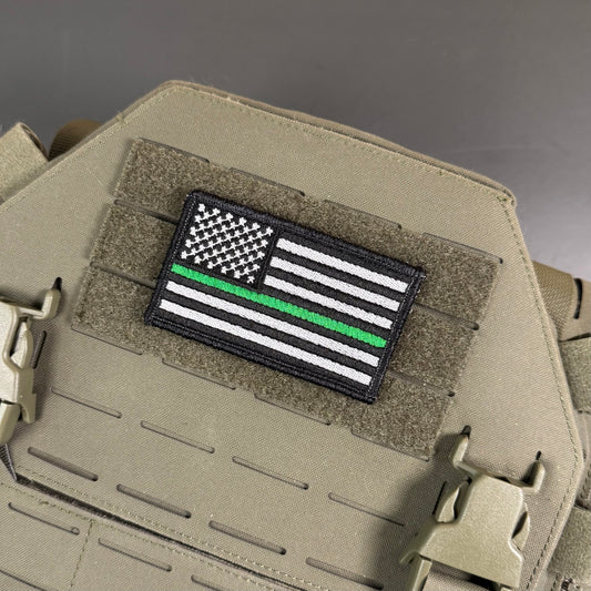 A rectangular embroidered patch featuring a black-and-white American flag with a single green stripe, mounted on tan tactical gear with Velcro and plastic clips.