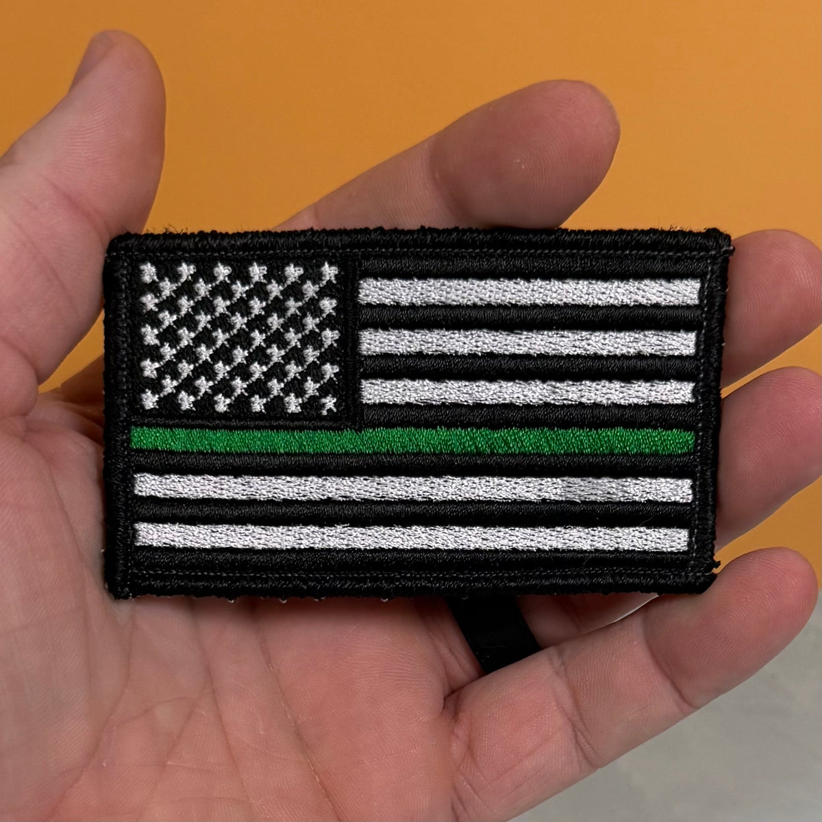 Thin Green Line Tactical American Flag embroidered morale patch: featuring a black-and-white American flag with a single green stripe, held in a person's hand against an orange background.