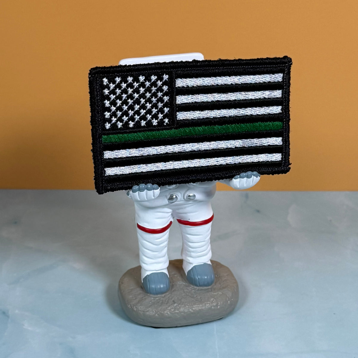 Thin Green Line Tactical American Flag embroidered morale patch: small astronaut figurine holding a rectangular embroidered patch with a black-and-white American flag design and a single green stripe, standing on a gray base against an.