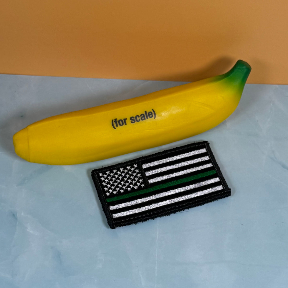 Thin Green Line Tactical American Flag embroidered morale patch: yellow banana-shaped item with green ends and the text '(for scale)' next to a black-and-white American flag patch with a thin green line, shown on a light blue surface with an.