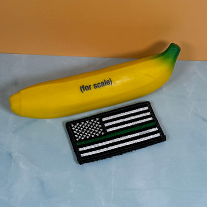 Thin Green Line Tactical American Flag embroidered morale patch: yellow banana-shaped item with green ends and the text '(for scale)' next to a black-and-white American flag patch with a thin green line, shown on a light blue surface with an.