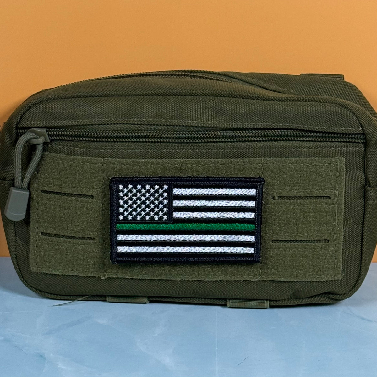 Thin Green Line Tactical American Flag embroidered morale patch: olive green tactical pouch with a front zipper and a rectangular embroidered American flag patch featuring a thin green line, set against a split orange and blue background.