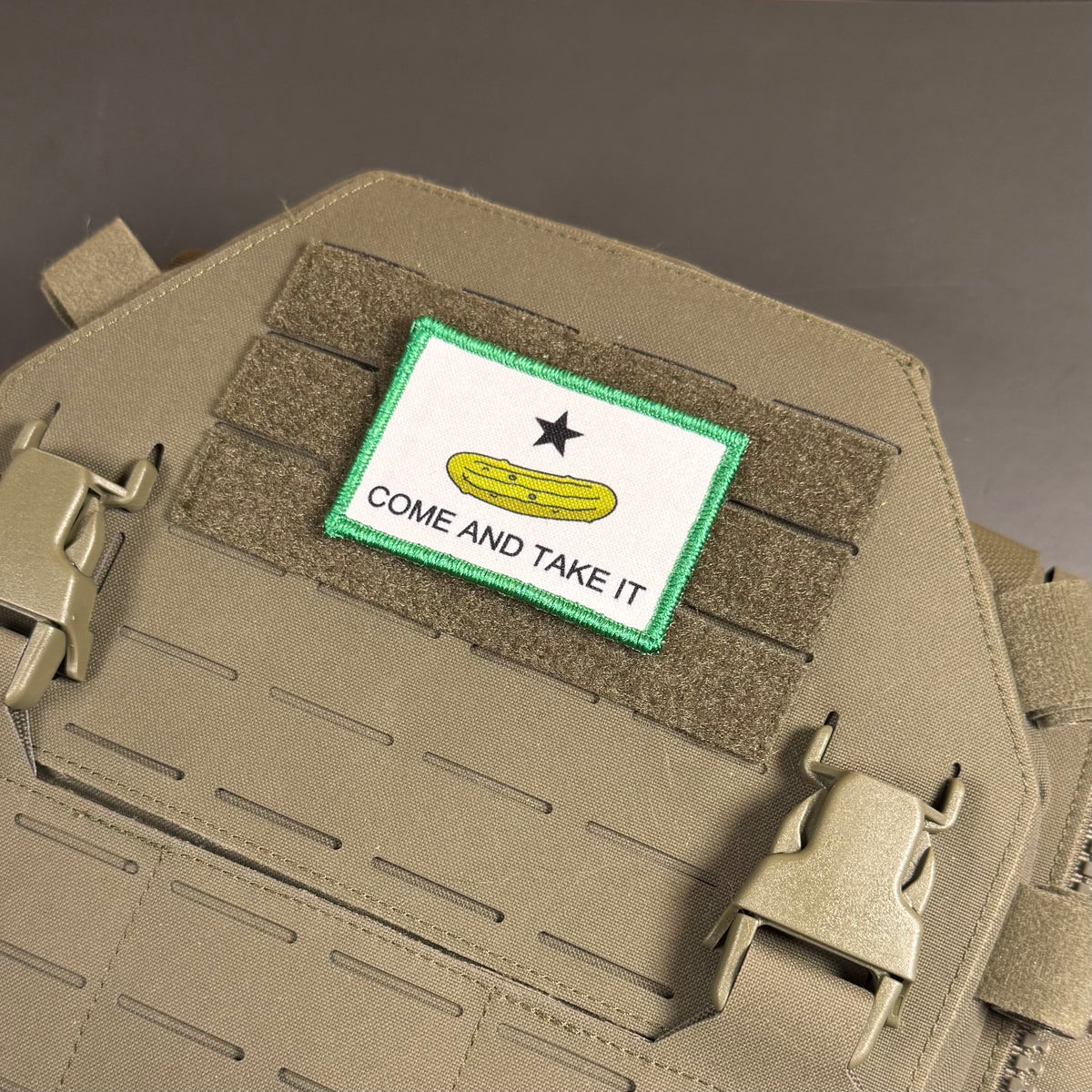 Come and Take It Pickle printed canvas morale patch, green border, black star, yellow pickle graphic, and the design lettering attached to tan tactical gear with velcro and quick-release buckles.