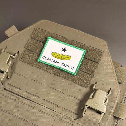 A rectangular embroidered patch with a white background, green border, black star, yellow pickle graphic, and the words 'COME AND TAKE IT' attached to tan tactical gear with velcro and quick-release buckles.