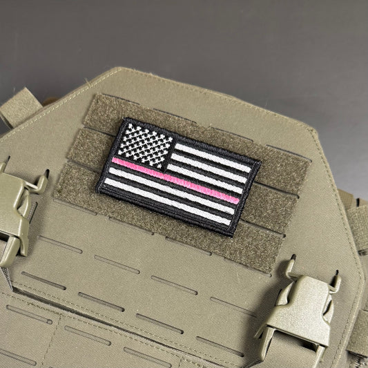 A rectangular embroidered American flag patch with a single pink stripe, displayed on a tan tactical vest with MOLLE webbing and quick-release buckles.