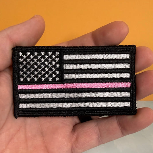 A hand holding a rectangular embroidered American flag patch with a single pink stripe across the middle, shown against an orange background.