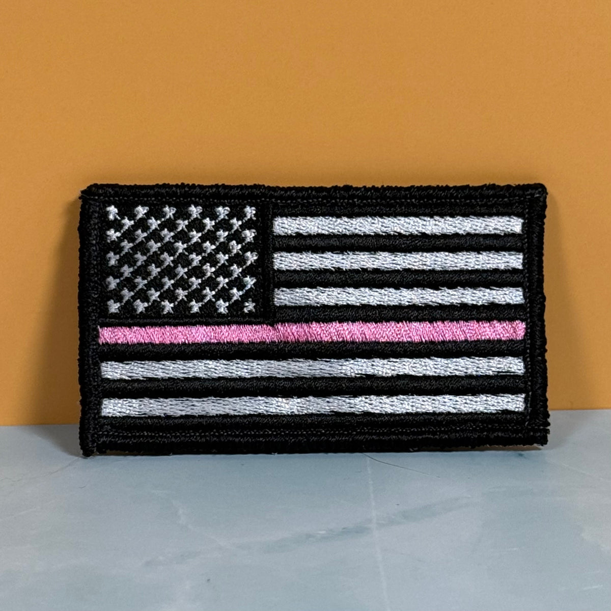 Breast Cancer Awareness Thin Pink Line American Flag embroidered morale patch: featuring a black-and-white American flag with a single bright pink horizontal stripe, photographed against a warm orange wall and light gray.