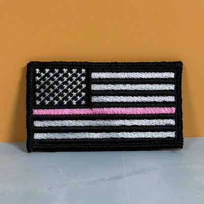 Breast Cancer Awareness Thin Pink Line American Flag embroidered morale patch: featuring a black-and-white American flag with a single bright pink horizontal stripe, photographed against a warm orange wall and light gray.