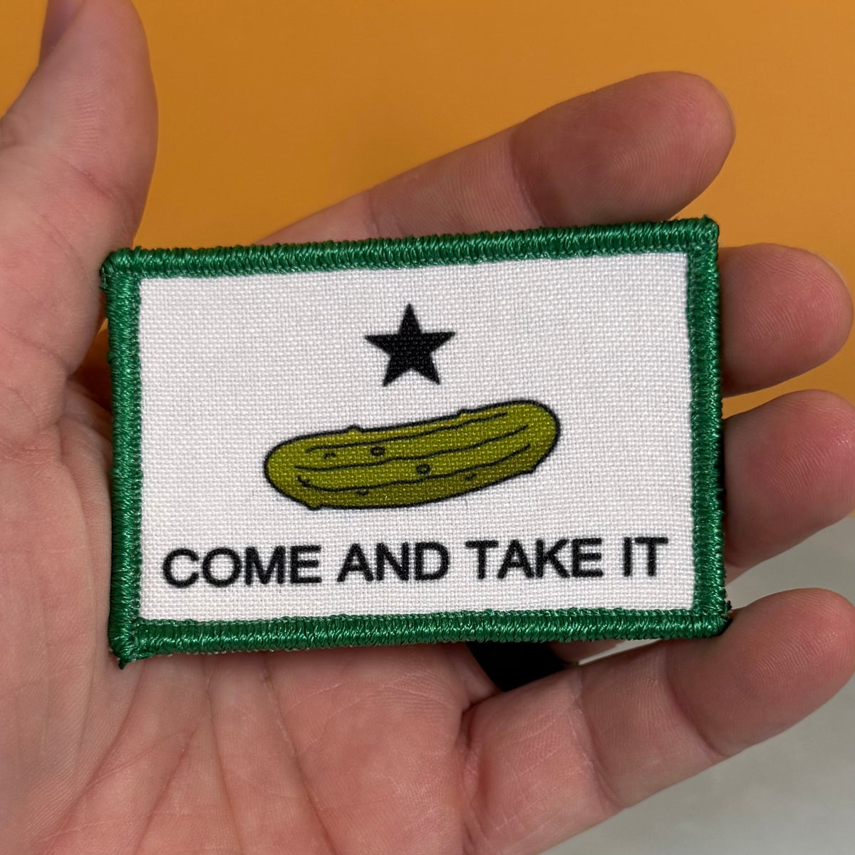 Come and Take It Pickle printed canvas morale patch, a hand holding a rectangular printed patch featuring a white field with a green border, a black star, a yellow-green pickle illustration, and the design lettering against an.
