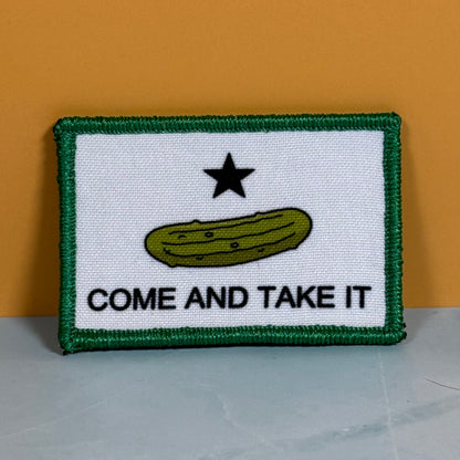 Come and Take It Pickle printed canvas morale patch featuring a green border, a white center, a black star, a green pickle illustration, and the design lettering in black, displayed against a solid orange background.