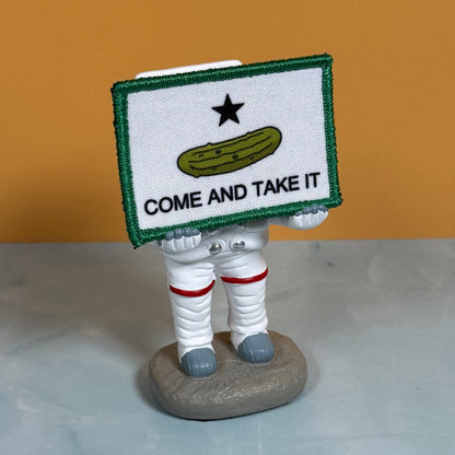 Come and Take It Pickle printed canvas morale patch, an astronaut figurine holding a rectangular printed patch with a green border, white background, black star, green pickle illustration, and the design lettering in black.