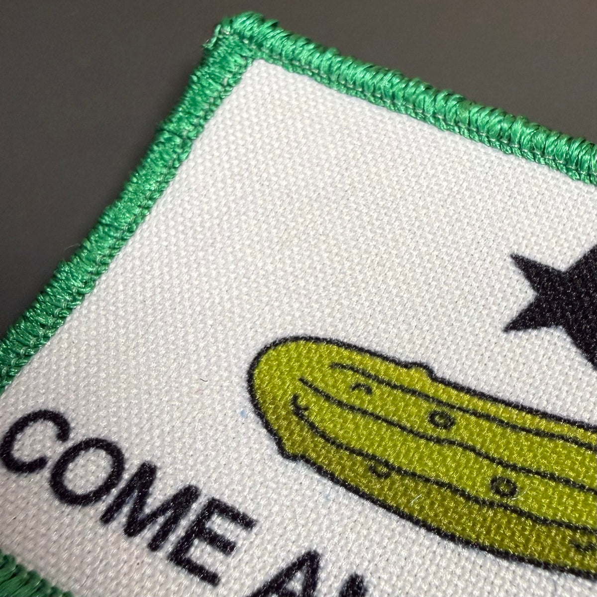 Come and Take It Pickle printed canvas morale patch, close-up of a square printed patch with a white field, green border, a yellow-green pickle illustration, a black star, and partial black text on a dark gray background.