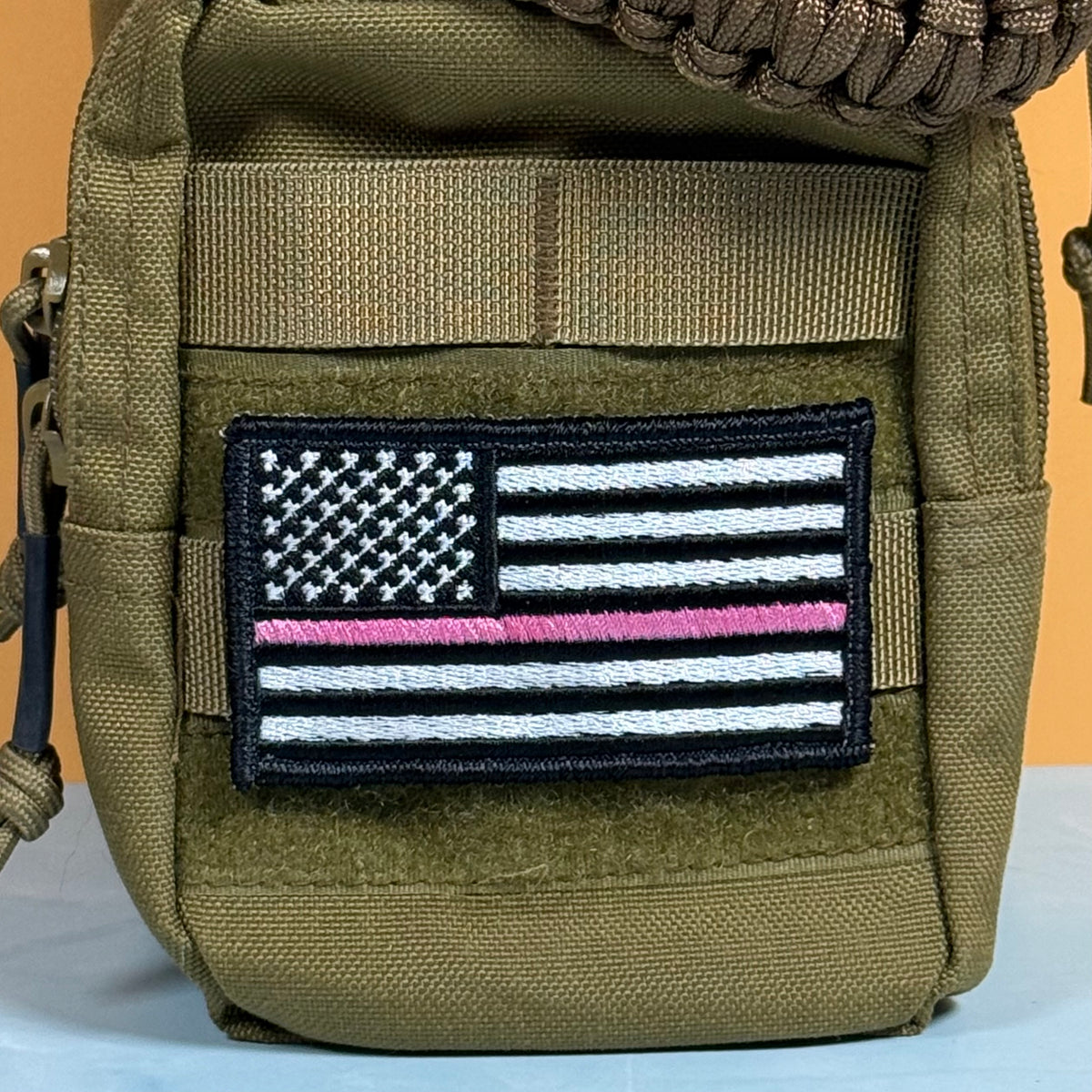 Breast Cancer Awareness Thin Pink Line American Flag embroidered morale patch: olive drab tactical pouch featuring a black, white, and pink embroidered American flag patch on the front, set against a solid orange background.