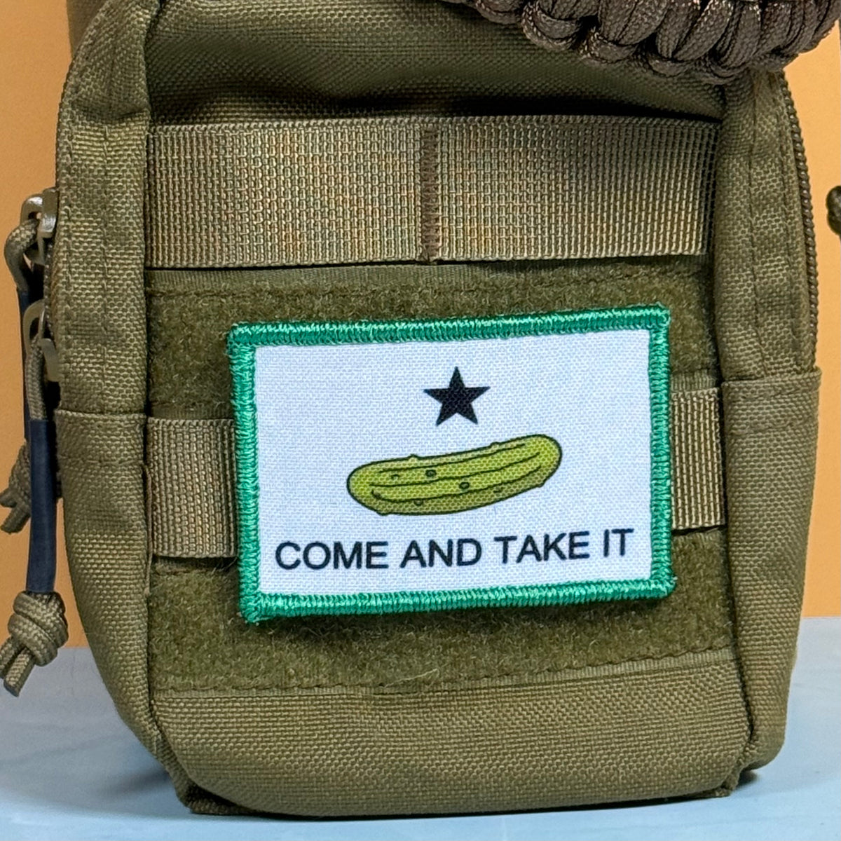 Come and Take It Pickle printed canvas morale patch, an olive drab tactical pouch featuring a custom printed patch with a white background, a green pickle, a black star, and the design lettering on a tan background.