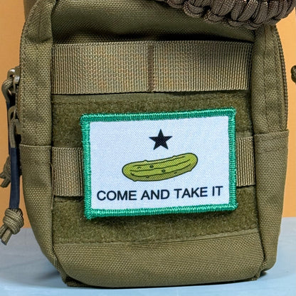 Come and Take It Pickle printed canvas morale patch, an olive drab tactical pouch featuring a custom printed patch with a white background, a green pickle, a black star, and the design lettering on a tan background.