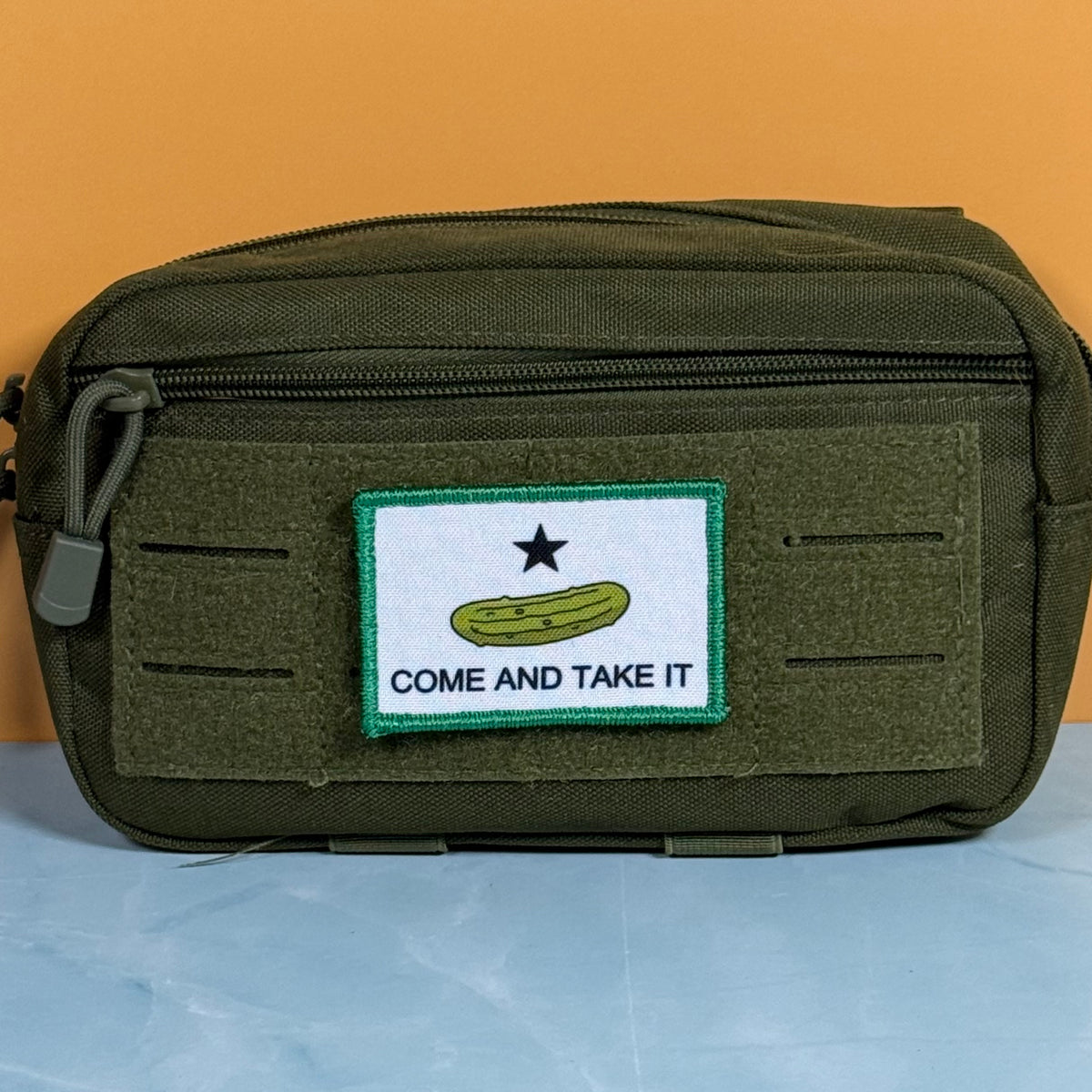 Come and Take It Pickle printed canvas morale patch, an olive green tactical pouch with a front zippered pocket and a rectangular 'Come and Take It' patch, set against a split orange and light blue background.