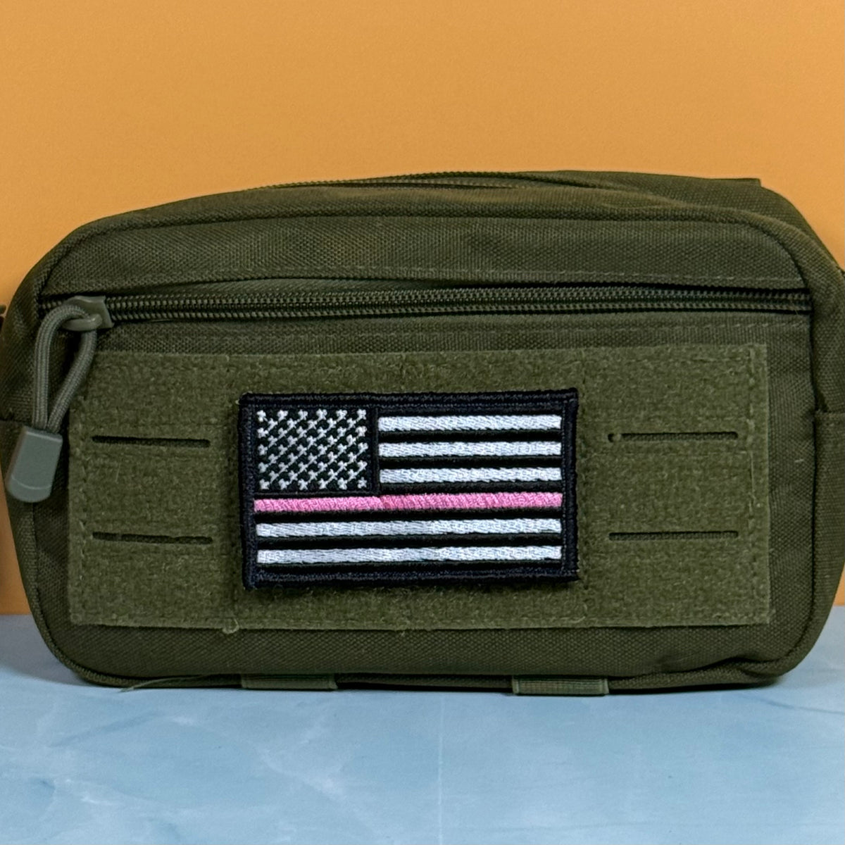 Breast Cancer Awareness Thin Pink Line American Flag embroidered morale patch: olive green tactical pouch with a front zippered pocket and a rectangular embroidered American flag patch featuring a single pink stripe, shown against an orange.