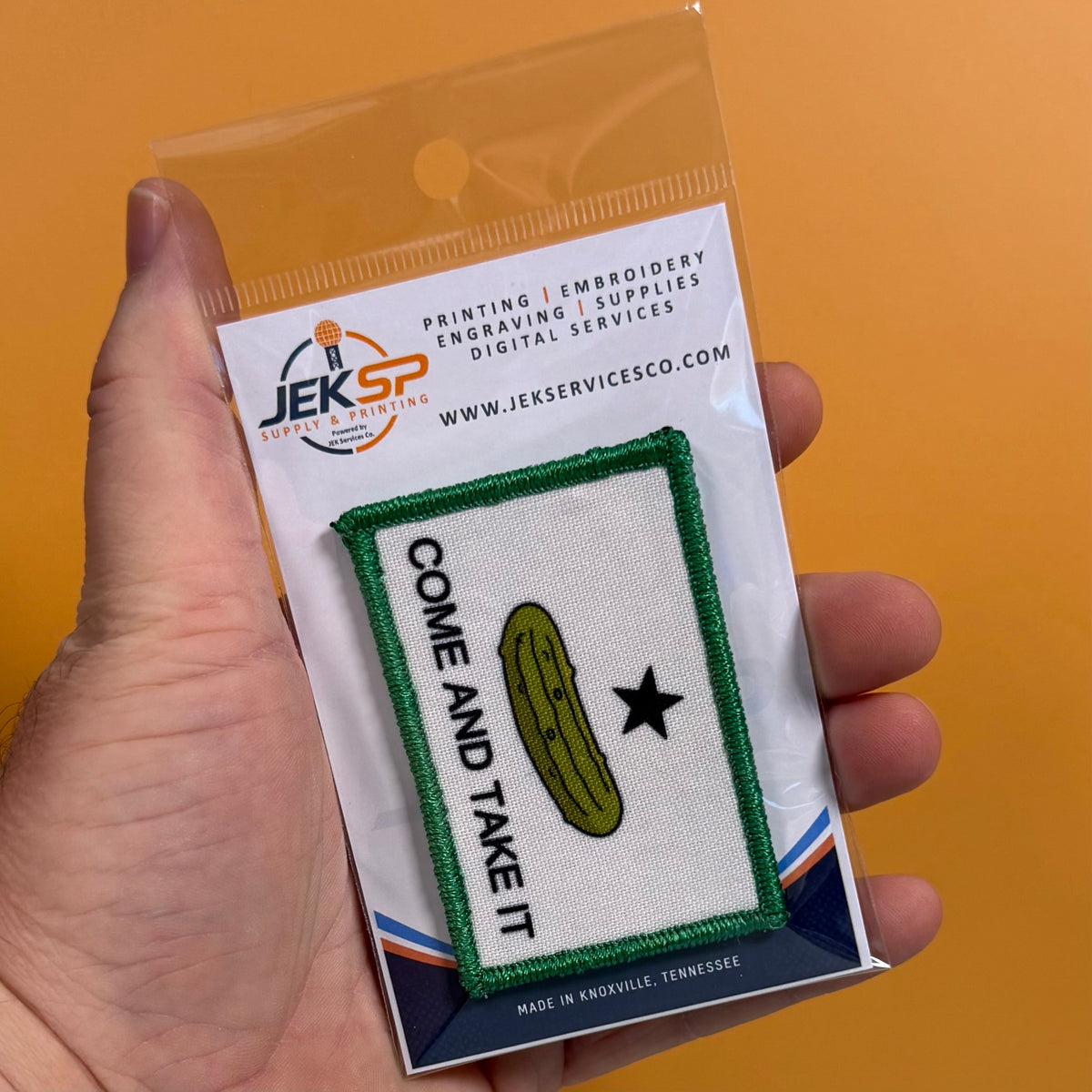 Come and Take It Pickle printed canvas morale patch, a hand holds a packaged printed patch featuring a green border, a yellow-green pickle illustration, a black star, and the design lettering on a white backing card. The.