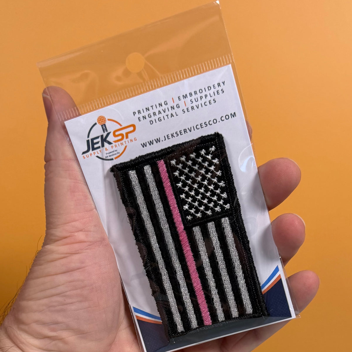 Breast Cancer Awareness Thin Pink Line American Flag embroidered morale patch: hand holds a packaged embroidered patch featuring a black-and-white American flag with a single pink stripe, on a white backing card. The card shows the JEKSP.