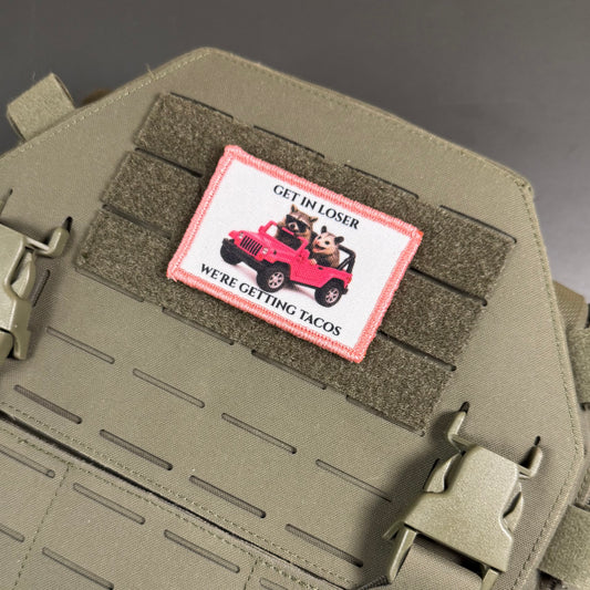 A rectangular embroidered patch with a pink border and a cartoon dog in a red jeep, reading 'GET IN LOSER WE'RE GETTING TACOS', attached to olive drab tactical webbing on a matching olive drab bag.