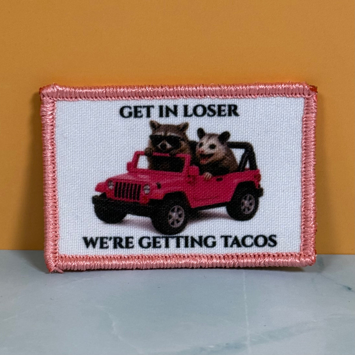 Get In Loser We're Getting Tacos printed canvas morale patch, white background, featuring a red jeep with a raccoon and opossum inside, and the design letteringRE GETTING TACOS'. The patch is displayed against a solid mustard.