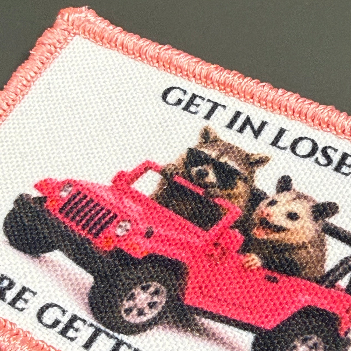 Get In Loser We're Getting Tacos printed canvas morale patch, a close-up of a rectangular printed patch with a pink stitched border, featuring two raccoons in a red jeep with partial text the design lettering and 'RE GETTE'.