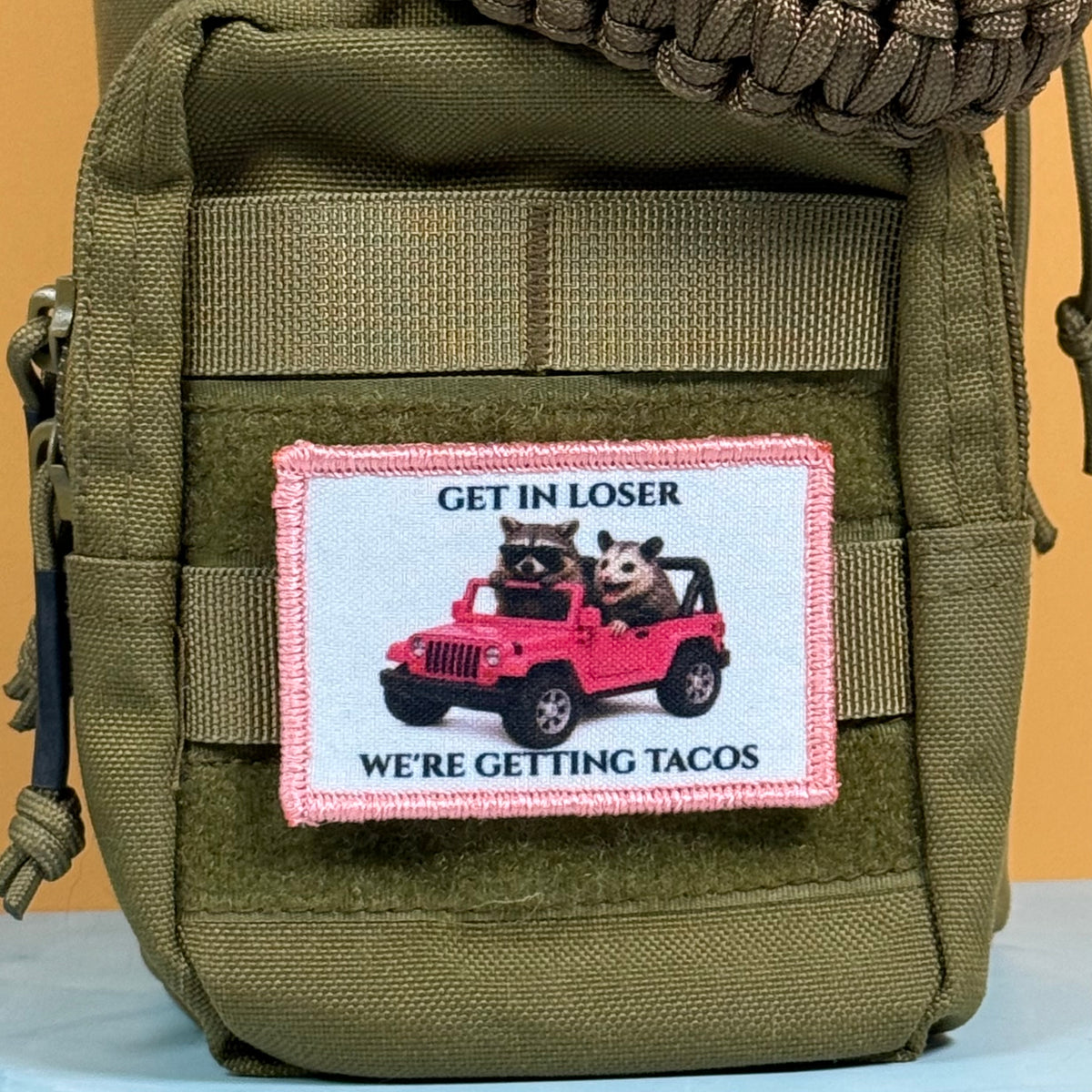 Get In Loser We're Getting Tacos printed canvas morale patch, an olive green tactical pouch featuring a rectangular printed patch with a pink border. The patch displays a cartoon raccoon and a cat driving a pink jeep, with the.