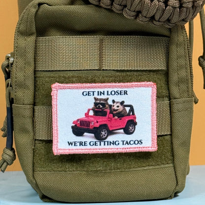 Get In Loser We're Getting Tacos printed canvas morale patch, an olive green tactical pouch featuring a rectangular printed patch with a pink border. The patch displays a cartoon raccoon and a cat driving a pink jeep, with the.