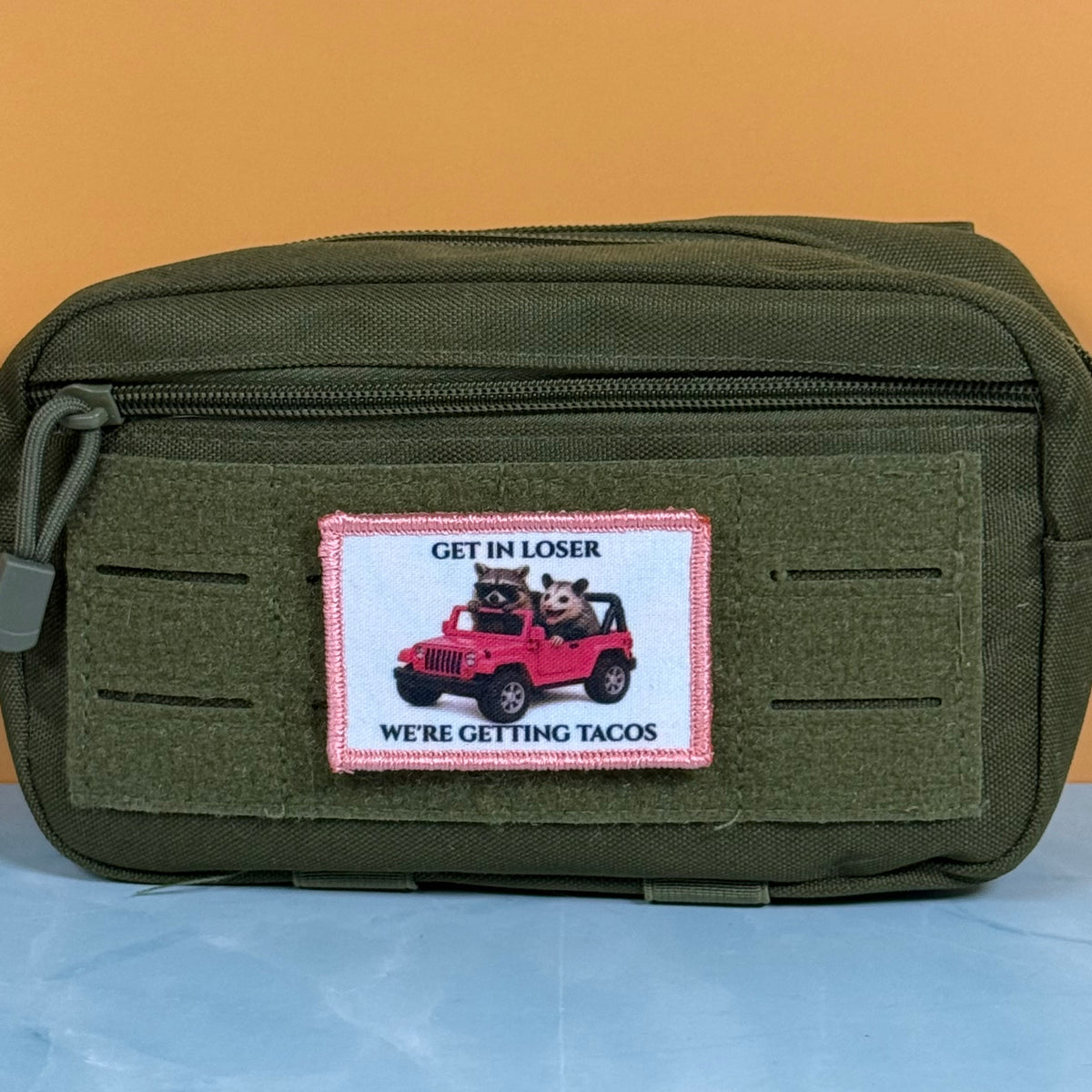 Get In Loser We're Getting Tacos printed canvas morale patch, an olive green canvas pouch with a front zipper and a rectangular patch featuring a red jeep with two dogs and the design letteringRE GETTING TACOS'.