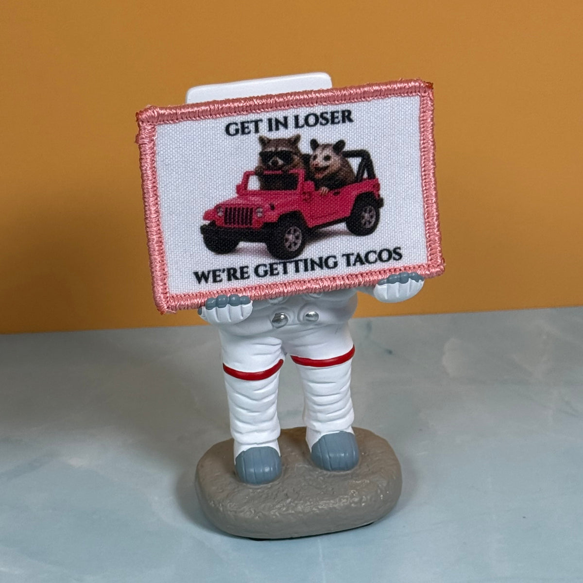Get In Loser We're Getting Tacos printed canvas morale patch, a novelty astronaut figurine holding a rectangular printed patch that the design letteringRE GETTING TACOS' with a pink jeep and two raccoons. The astronaut is.