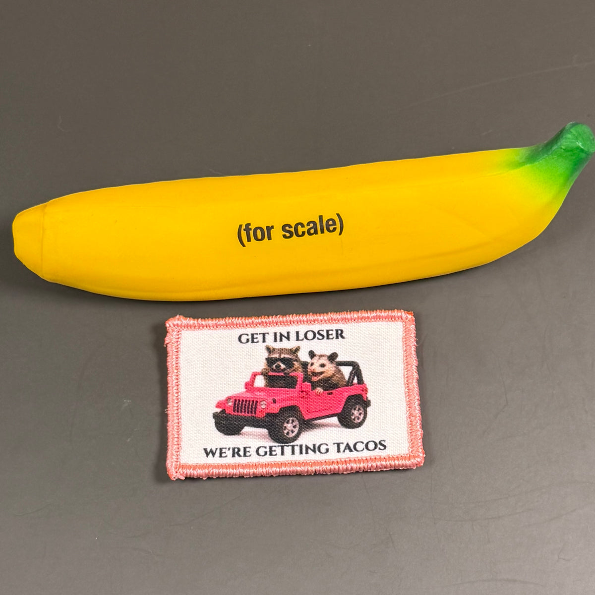 Get In Loser We're Getting Tacos printed canvas morale patch, a yellow novelty item shaped like a banana with green tips and the design lettering above it, shown next to a small rectangular patch with a raccoon in a pink jeep.