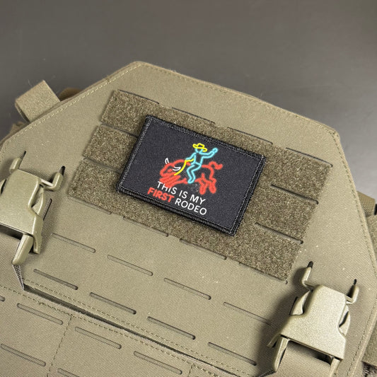 A close-up of a tactical vest or plate carrier in olive drab tan, featuring a black rectangular embroidered patch with a colorful stick-figure rider and the text 'THIS IS MY FIRST RODEO'.