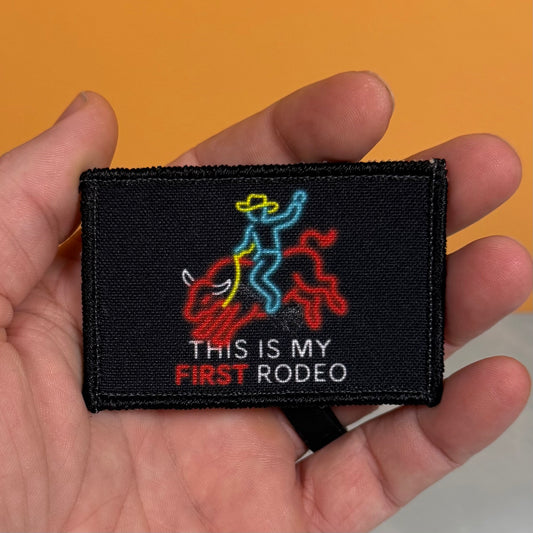A hand holding a rectangular black embroidered patch with a cowboy riding a bull graphic and the text 'THIS IS MY FIRST RODEO' against a solid orange background.
