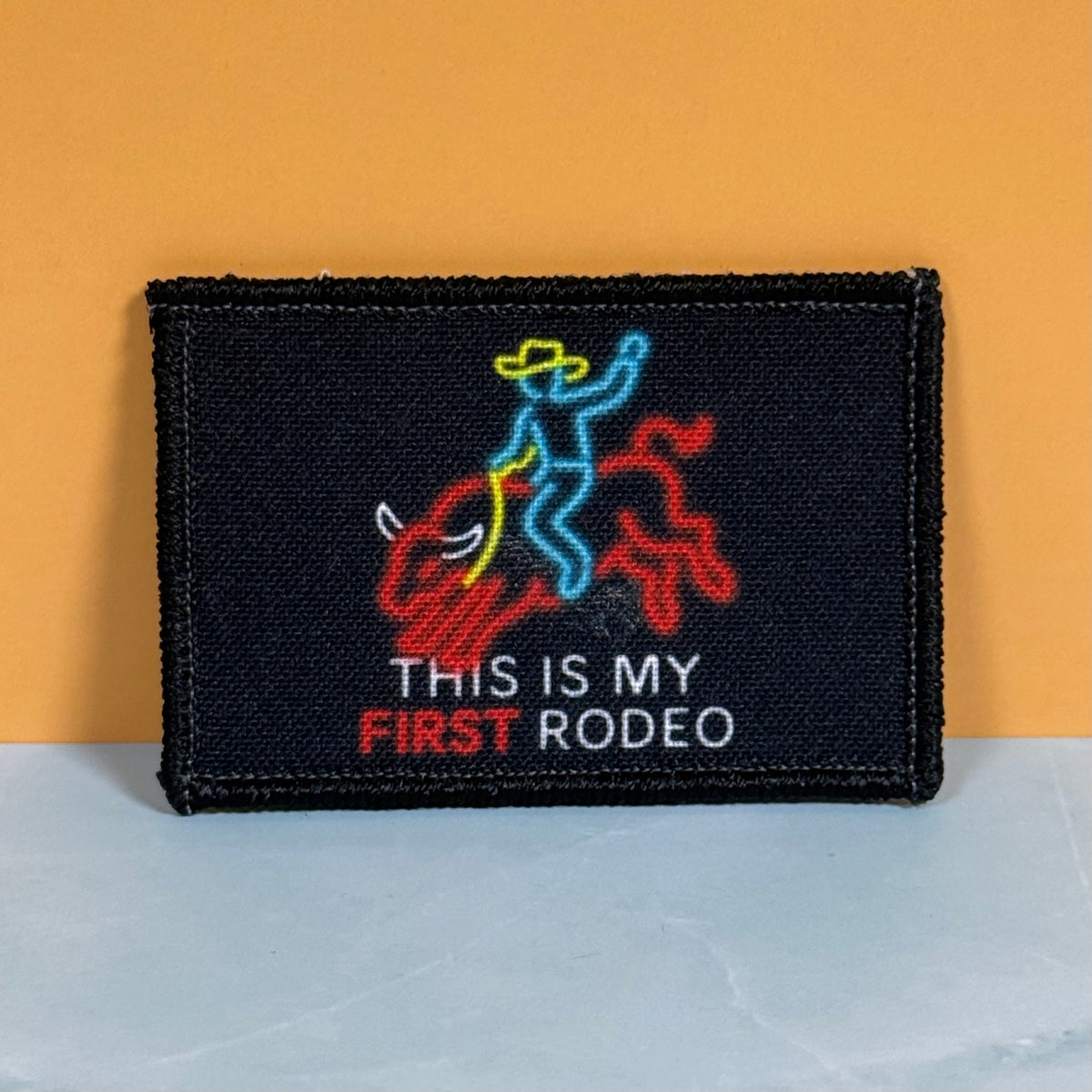 My First Rodeo printed canvas morale patch featuring a neon-style cowboy riding a red bull, with the design lettering in white and red. The patch is photographed standing upright against a two-tone background with a mustard.