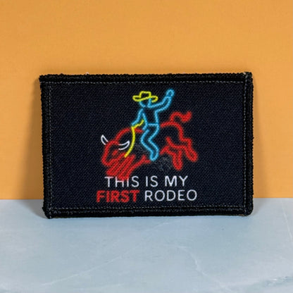 My First Rodeo printed canvas morale patch featuring a neon-style cowboy riding a red bull, with the design lettering in white and red. The patch is photographed standing upright against a two-tone background with a mustard.