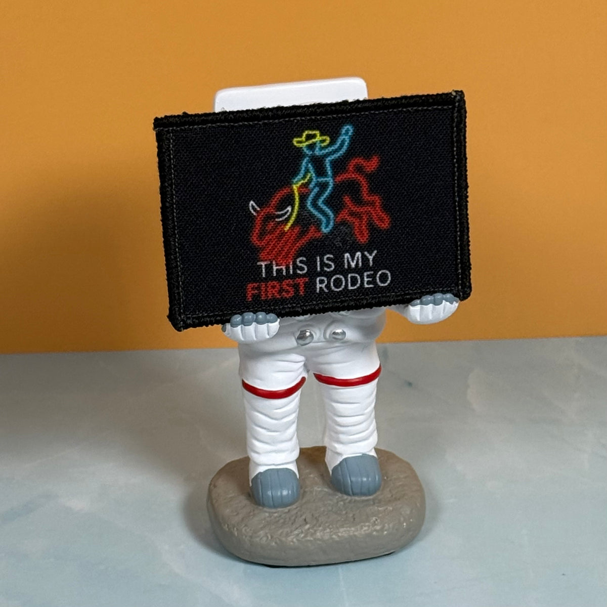My First Rodeo printed canvas morale patch, a small astronaut figurine holding a black rectangular patch that the design lettering with a colorful cowboy-and-rider graphic. The astronaut is white with red stripes and gray.