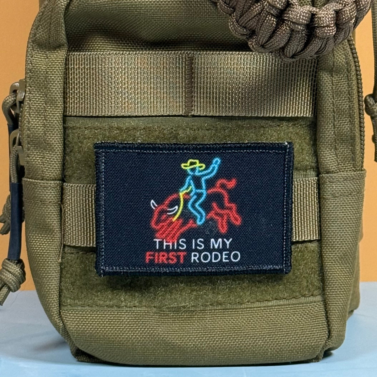 My First Rodeo printed canvas morale patch, an olive green tactical pouch with a black rectangular patch that the design lettering and features a colorful neon-style rider and horse graphic, shown against a simple background.