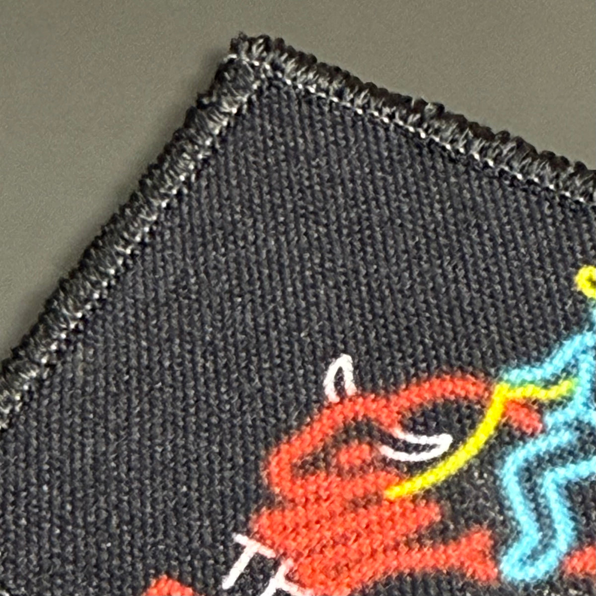 My First Rodeo printed canvas morale patch, close-up of a dark woven fabric patch with a colorful printed design, showing a red figure, a yellow curved line, and a light blue outlined figure on a dark charcoal background.