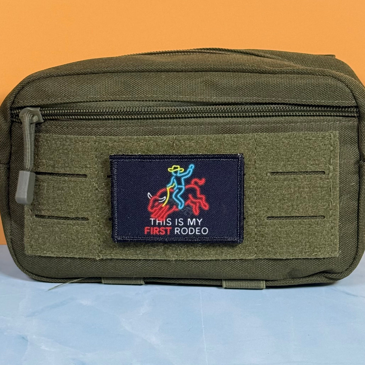 My First Rodeo printed canvas morale patch, an olive green tactical pouch with a front zipper and a central printed patch that the design lettering with a cowboy boot and red bull graphic, set against a split orange and light.