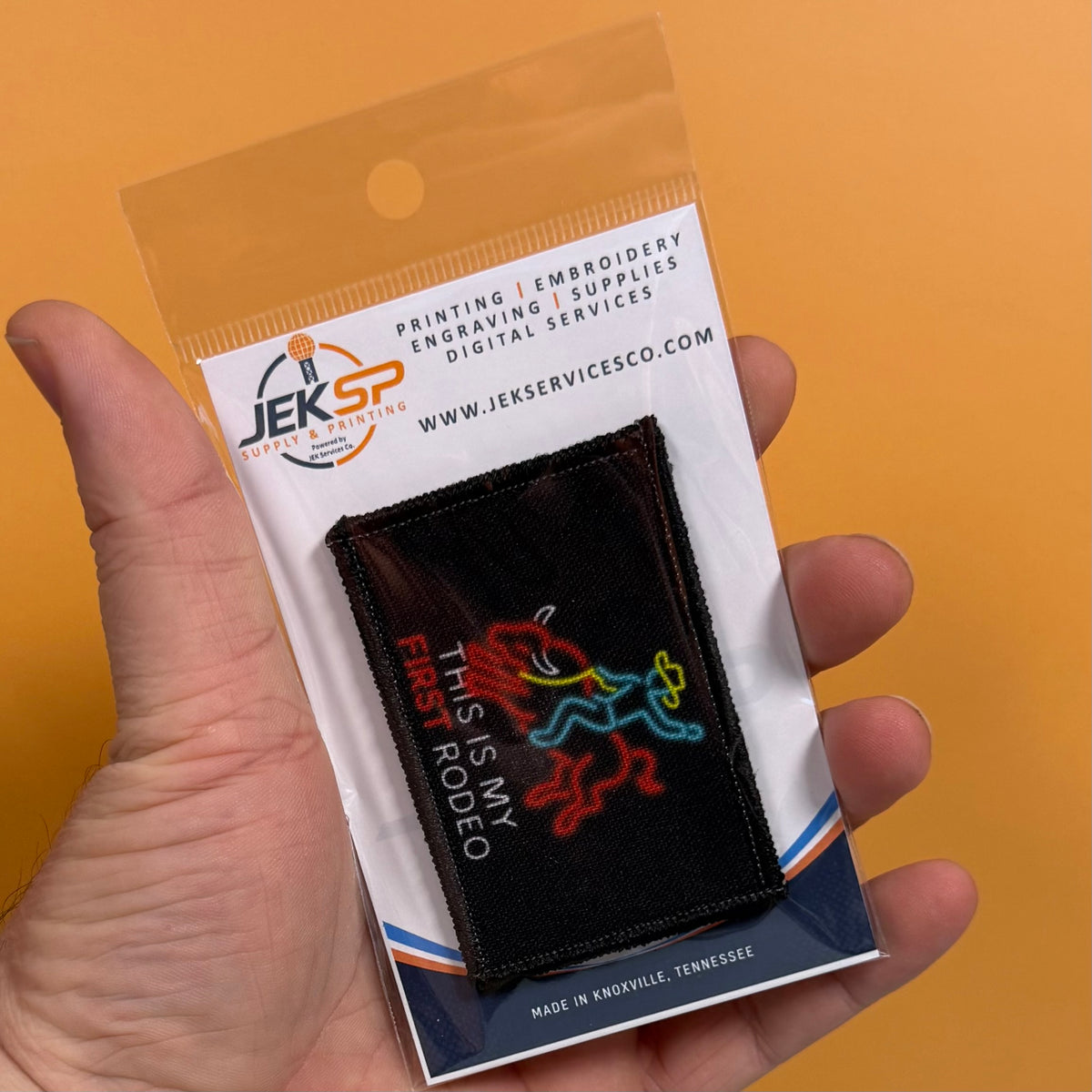My First Rodeo printed canvas morale patch, a hand holds a packaged printed patch against a solid orange background. The patch is black with a colorful neon-style horse and rider design and the design lettering.