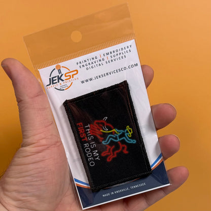 My First Rodeo printed canvas morale patch, a hand holds a packaged printed patch against a solid orange background. The patch is black with a colorful neon-style horse and rider design and the design lettering.