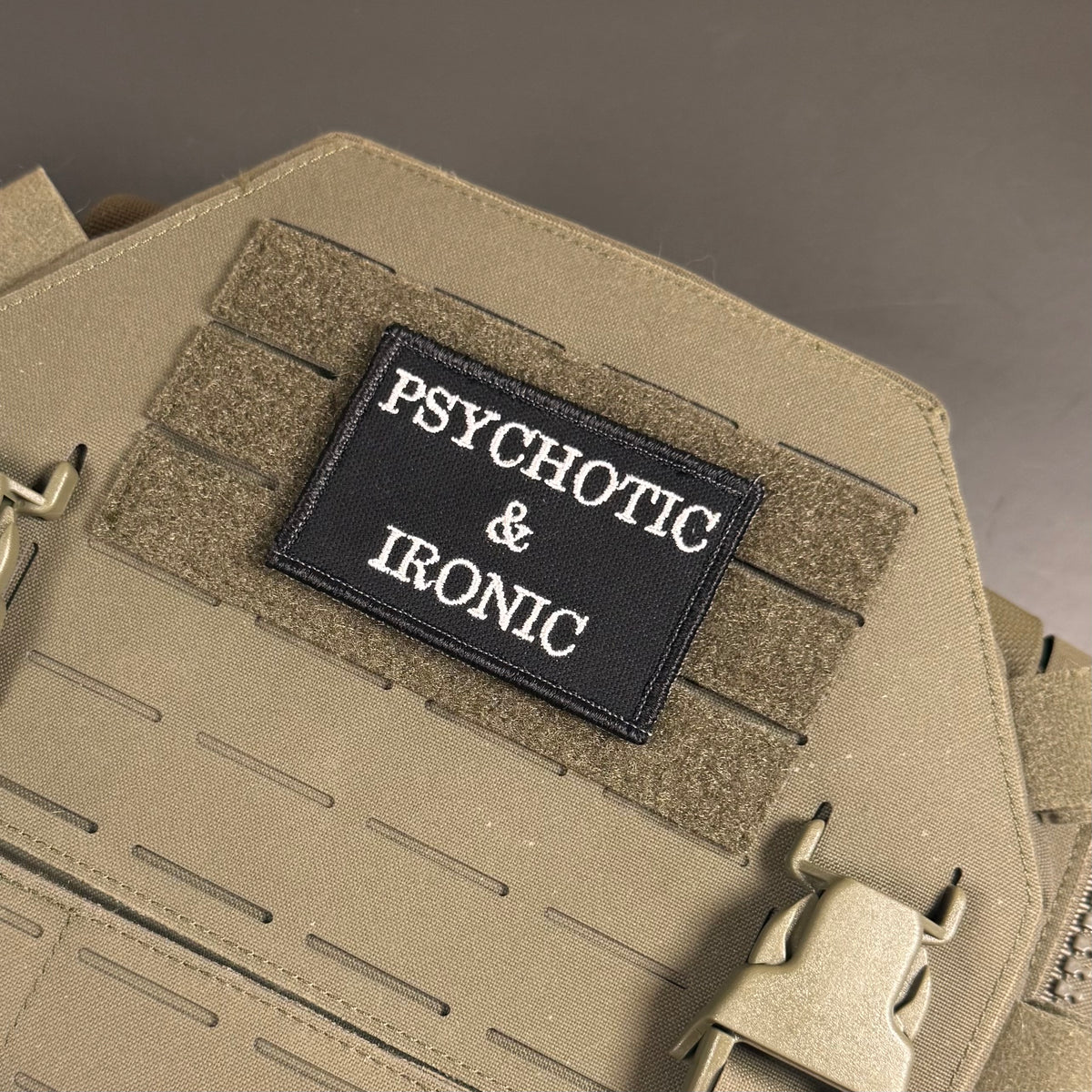 A close-up of a tactical vest or backpack in coyote tan with a black rectangular patch reading 'PSYCHOTIC & IRONIC' in white text, attached to Velcro strips.