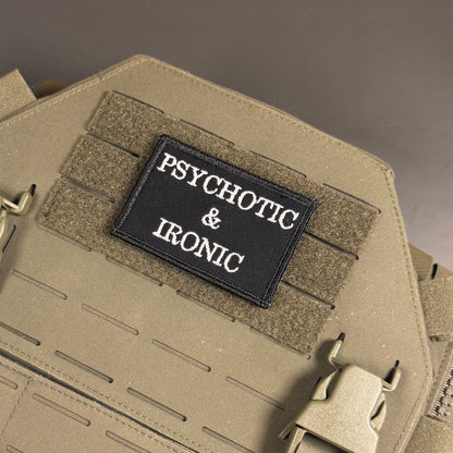 A close-up of a tactical vest or backpack in coyote tan with a black rectangular patch reading 'PSYCHOTIC & IRONIC' in white text, attached to Velcro strips.